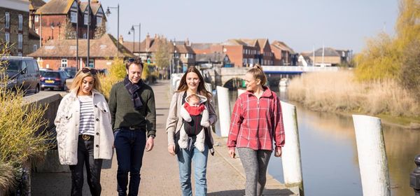 Three women and one man walking towards the camera along the quay at Sandwich next to the river with buildings in the background