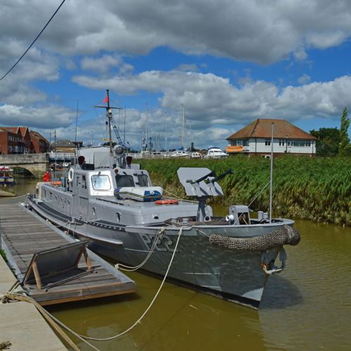 A grey patrol boat on the river next to a boarding quay with buildings in the distance