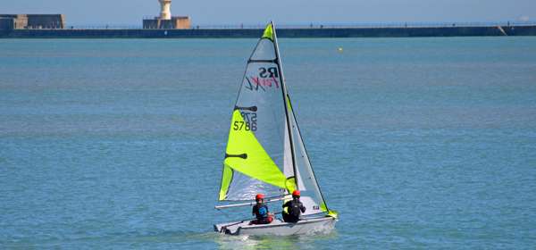 Two people wearing red protective helmets on a sailing dingy with a fluorescent yellow sail in Dover Harbour.