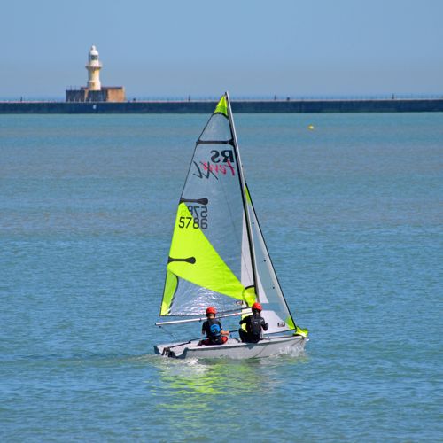 Two people wearing red protective helmets on a sailing dingy with a fluorescent yellow sail in Dover Harbour.