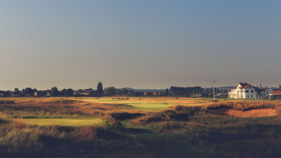 A white clubhouse to the right of the image with a golf green in front, rough and bunkers. 