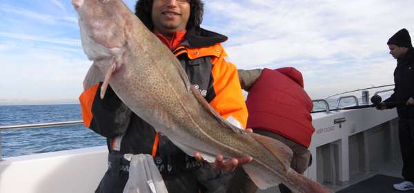 A man wearing a black and orange jacket and a fur hat holding a large fish aboard a fishing boat at sea.