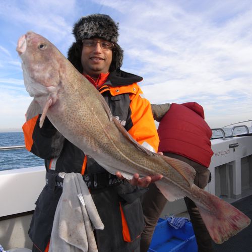 A man wearing a black and orange jacket and a fur hat holding a large fish aboard a fishing boat at sea.