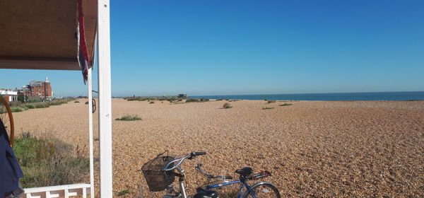 Two bicycles parked up next to a beach hut on a shingle beach with the sea and blue sky in the distance. 