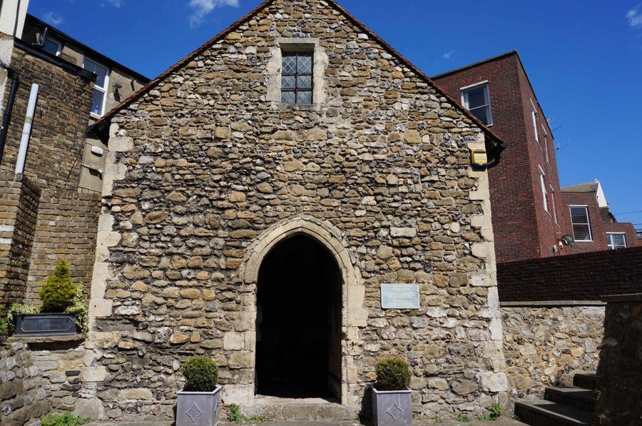 The stone facade of tiny St Edmund's Chapel in Dover flanked by modern buildings.
