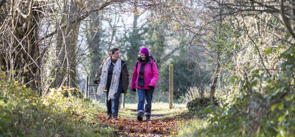 Two women walking along a woodland path, one wearing a pink coat and hat, both wearing walking boots.