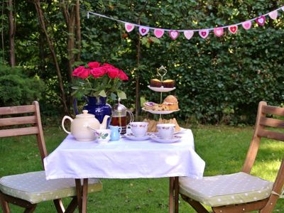 A table and two chairs outside laid with a white tablecloth and an afternoon tea for two with bunting in the background.