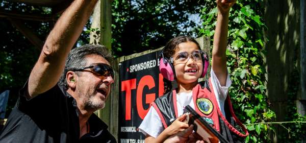 A man wearing protective goggles instructing a young girl holding a shotgun (also wearing protective goggles and pink ear protectors) in clay pigeon shooting.