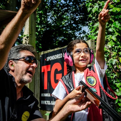 A man wearing protective goggles instructing a young girl holding a shotgun (also wearing protective goggles and pink ear protectors) in clay pigeon shooting.