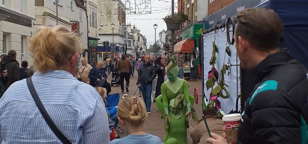 A busy high street with a street performer in a green outfit.