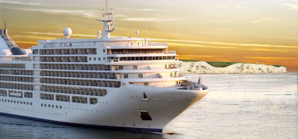 Large white cruise ship on a calm sea with the White Cliffs in the background and a yellow tint in the sky.