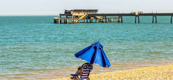 A blue and white striped deckchair with blue parasol, on a shingle beach with the sea and pier