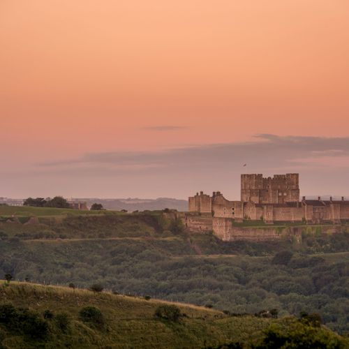 Dover Castle from a distance in the early morning light with a pink sky and greenery in the foreground.