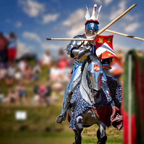 A knight on horseback riding towards the camera with jousting stick, crowds in the background.