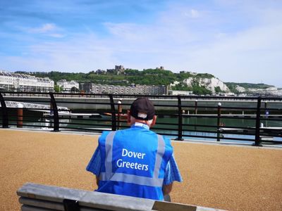 A man wearing a blue high-vis tabard with the words Dover Greeters on the back sitting on a bench in the foreground, with Dover seafront and castle in the distance