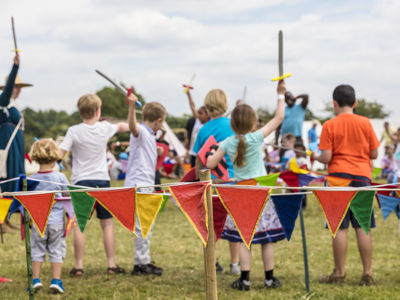 A group of children waving wooden swords facing away from the camera with multicoloured bunting in the foreground.