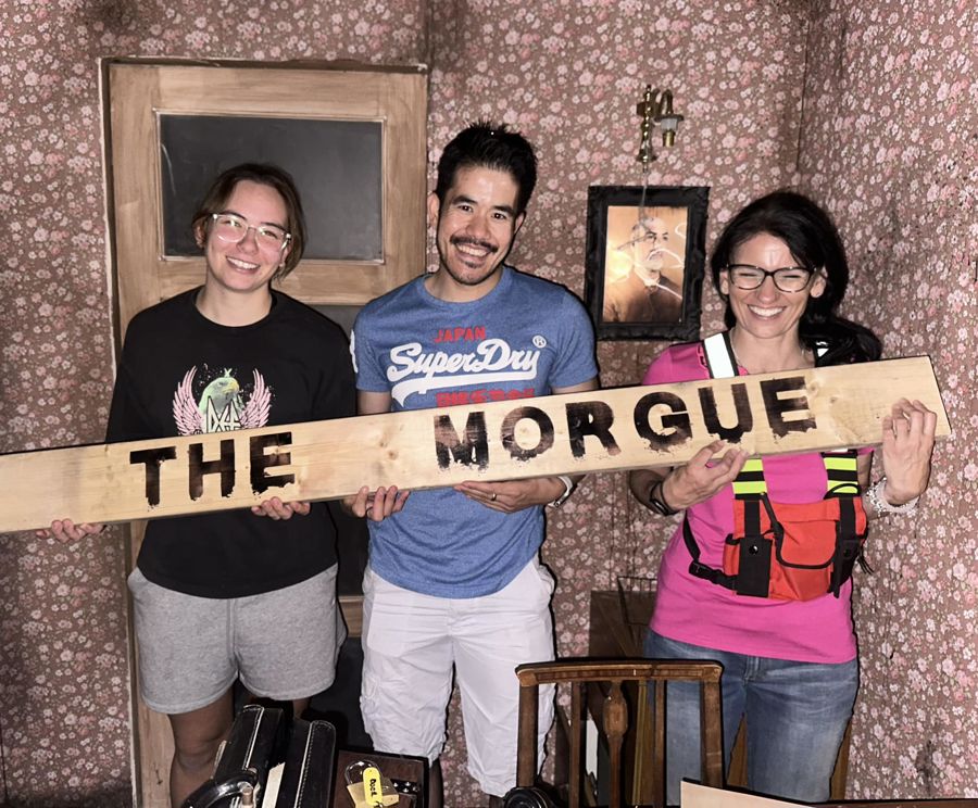 Three people in a room set holding a sign saying 'The Morgue'. 