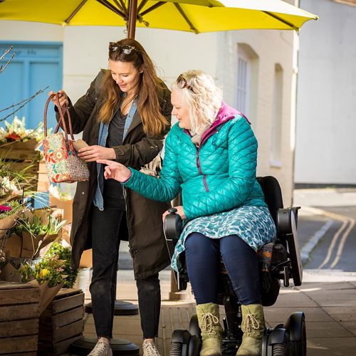 Two women, one in a wheelchair, under a yellow umbrella outside a flower shop.