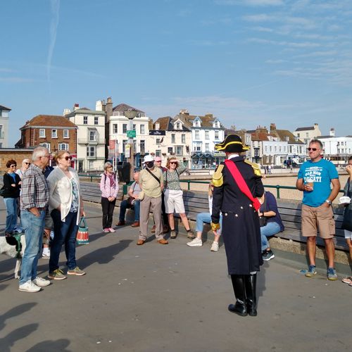 A man dressed in Napoleonic costume talking to a group of tourists on Deal Pier with the beach and seafront in the background.