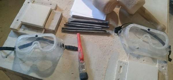 A wooden table with stone-carving tools laid out including mallets, chisels, goggles and stone blocks.