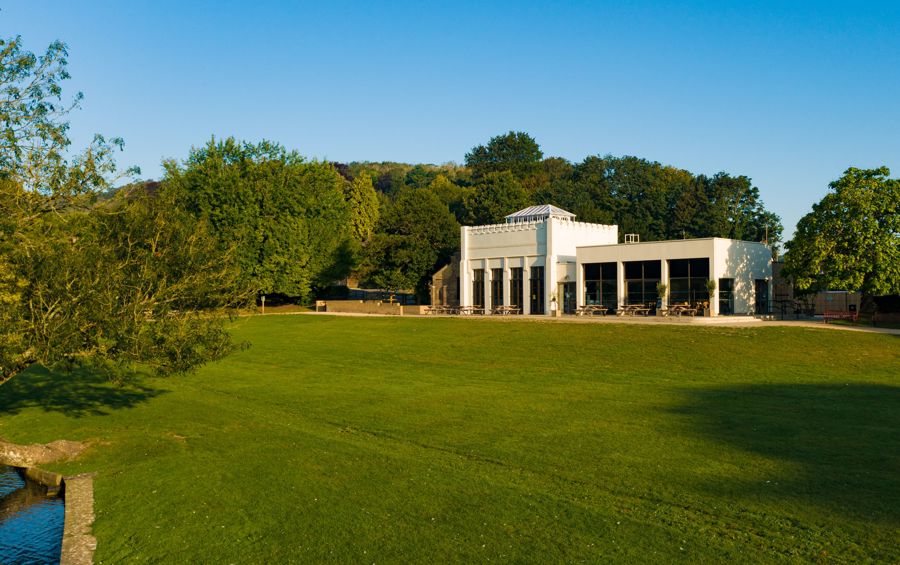 An area of green grass in front of a white building housing Kearsney Cafe with trees in the background and a blue sky.