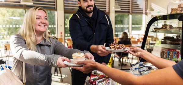 Two people at a cafe counter being handed cakes by the server.