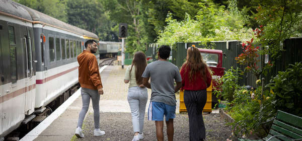 Four people on a station platform with a train to the left of the image and a green fence and trees to the right.