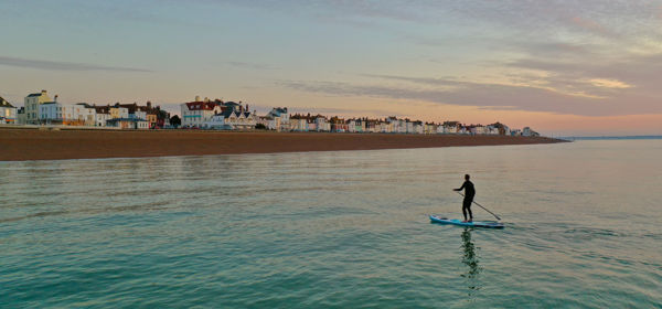 A person on a paddleboard on a calm sea with Deal seafront and beach in the background.