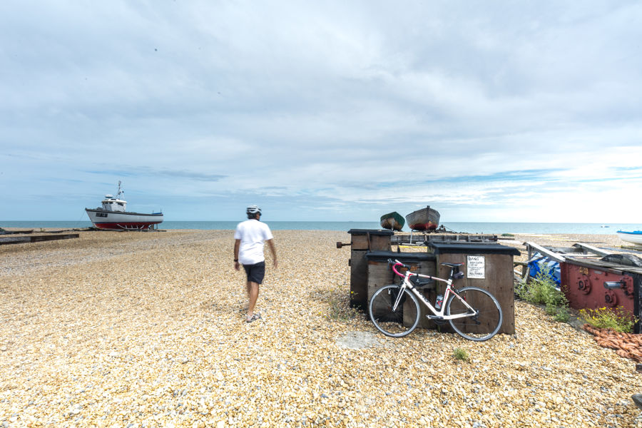 A bicycle on a shingle beach, with the rider walking towards the sea in the distance and a few boats.