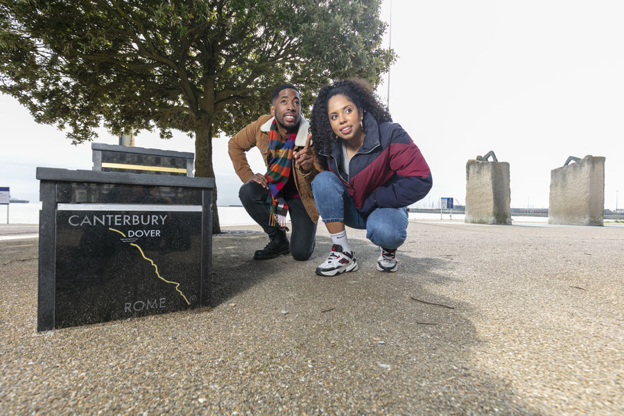 Two people crouched down beside a black granite sculpture of a winners podium on Dover seafront with the harbour in the background.