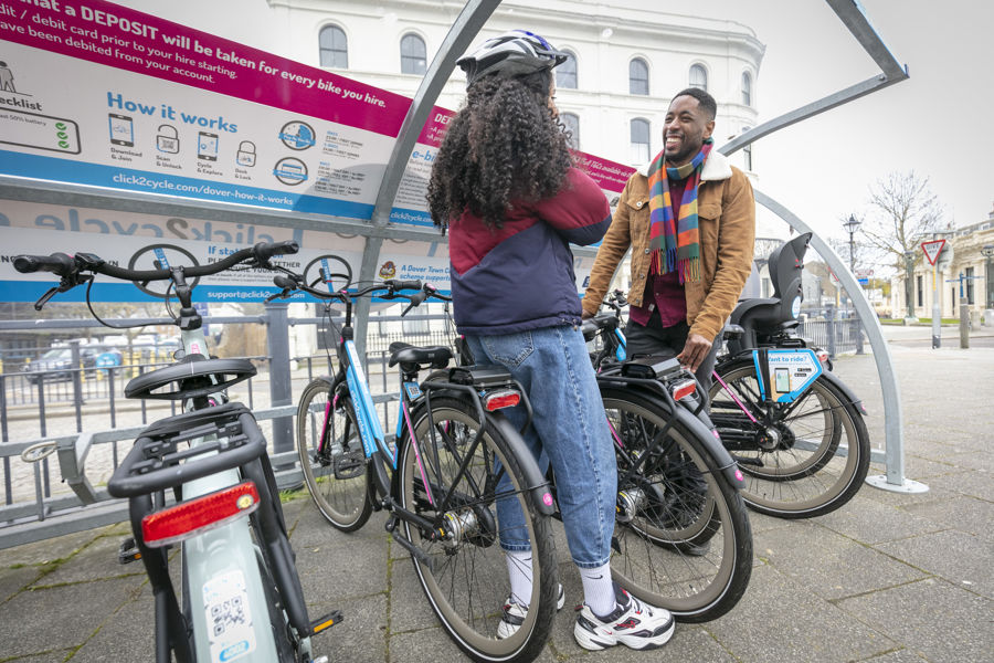 A woman putting on a bike helmet and a man holding a bike at a Click2Cycle bike station in Dover.