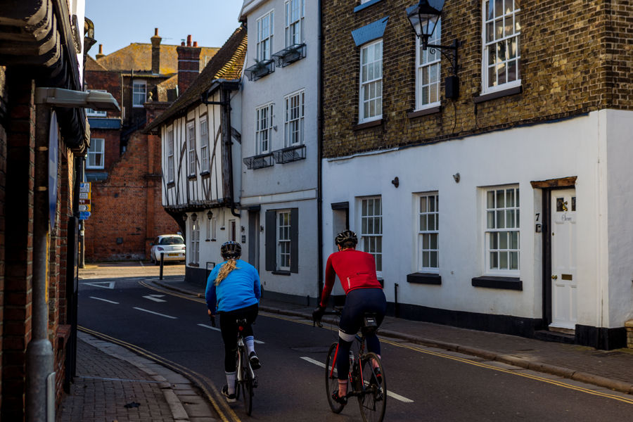 Two cyclists heading away from the camera along a street in Sandwich with medieval buildings on either side.