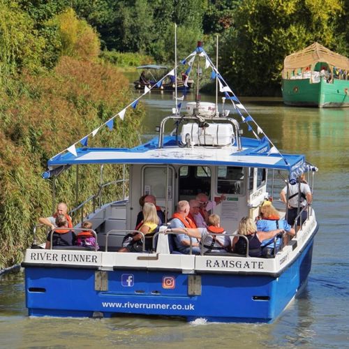 People aboard the blue and white Sandwich River Runner boat on the  River Stour heading away from the camera.