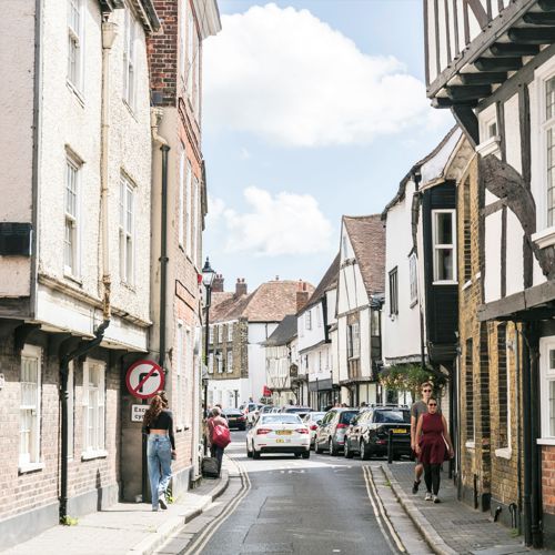 A street of medieval buildings in Sandwich a few people and cars in the distance.