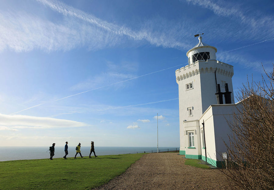 Four walkers walking on green grass from the left of the picture towards the white South Foreland Lighthouse on the right of the picture, on a winter's day, blue sky above.