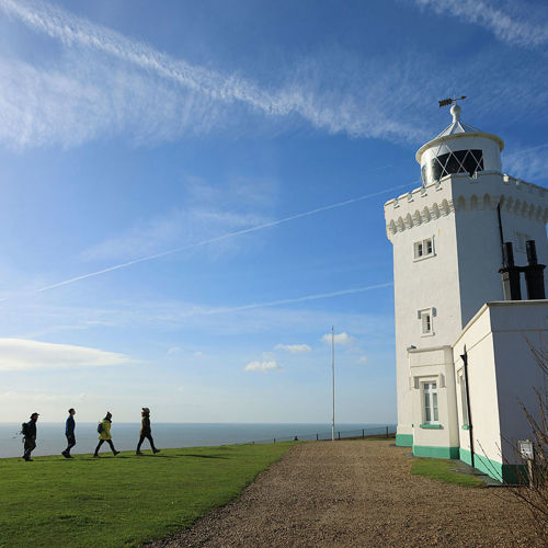 4 people approaching a white painted lighthouse on foot, across a green lawn with the sea just visible in the background.