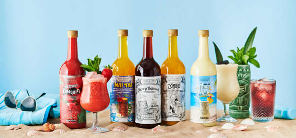 A selection of brightly coloured bottled cocktails and glasses with cocktails on sand with a towel and sunglasses to promote the summer cocktail range.