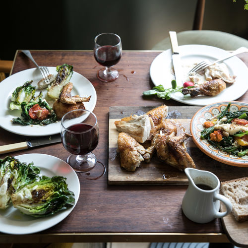 A dark wooden table with plates of food - roast chicken, vegetables and bread - and glasses of red wine.