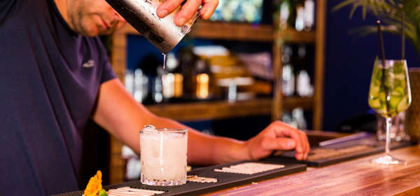 A man pouring a cocktail from a silver cocktail shaker into a glass on a wooden bar, bottles and glasses in the background.