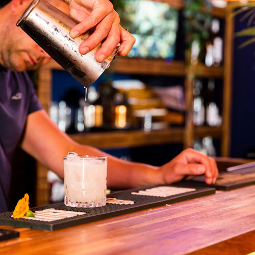 A man pouring a cocktail from a silver cocktail shaker into a glass on a wooden bar, bottles and glasses in the background.