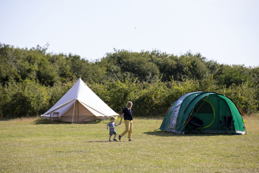 A woman and a small boy walking hand in hand across a grassy campsite with two tents in the background.