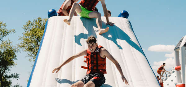 Young people wearing red life jackets and swimsuits sliding down a giant, floating, inflatable slide into blue water.